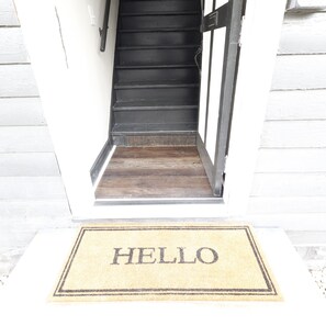 Eine Holztreppe führt zu einer Tür, vor der eine Fußmatte mit dem Schriftzug 'HELLO' liegt.