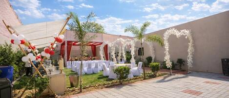 Un espacio para eventos al aire libre con mesas y sillas, un arco rojo y adornos florales blancos.
