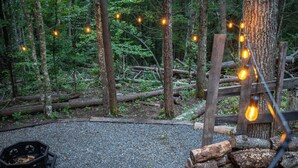 A forested area with a gravel path, wooden logs, and string lights. - Luxury Covered Wagon Stay • Firepit, Views, Stargazing (Sevierville)