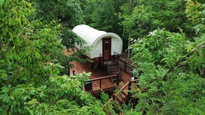 A white tent with a wooden door, surrounded by dense forest, connected to a wooden deck with a metal railing. - Luxury Covered Wagon Stay • Firepit, Views, Stargazing (Sevierville)
