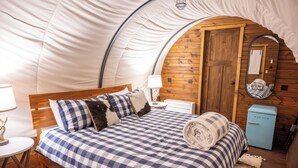 A tented room with a bed, a small table, a lamp, a mirror, and a blue refrigerator. - Luxury Covered Wagon Stay • Firepit, Views, Stargazing (Sevierville)