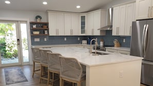 A modern kitchen with white cabinetry, a central island with bar stools, a stainless steel refrigerator, and a view of greenery outside. - Welcome to Hidden Oasis, your dog-friendly home away from home in Sanibel! (Sanibel)