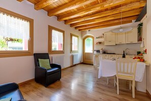Ein Zimmer mit Holzbalkendecke, einem schwarzen Ledersessel, einem Esstisch mit Stühlen und einer Küchenzeile mit Schränken und Herd.