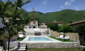 Ein Pool mit Liegestühlen, eine Steinmauer und Häuser in einer gebirgigen Gegend.