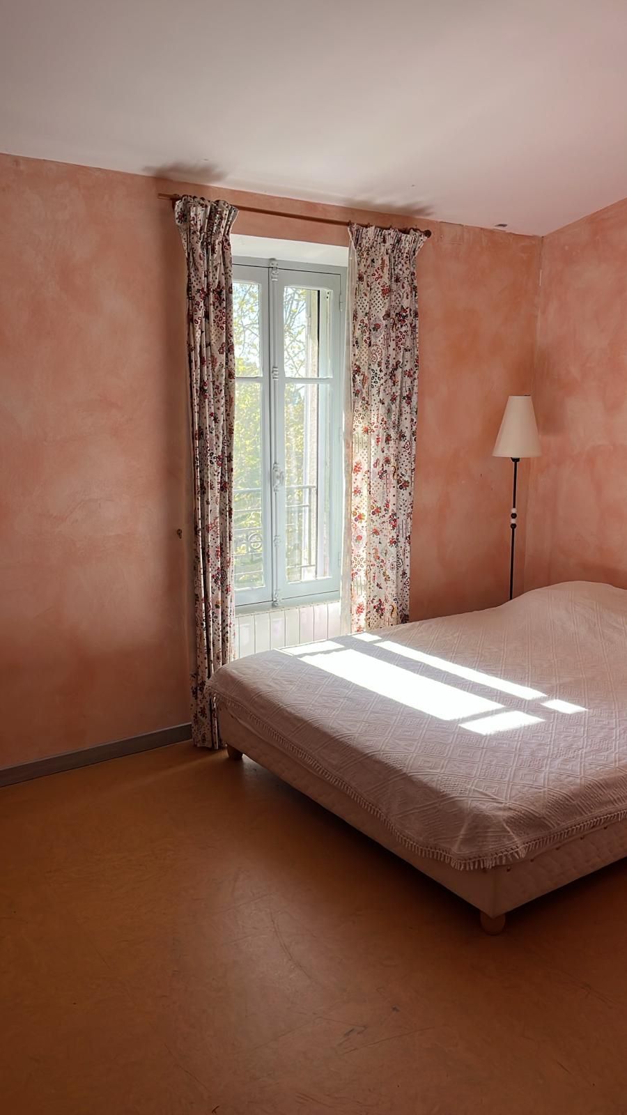 Een slaapkamer met een enkel bed, een raam met bloemen gordijnen en een vloerlamp.