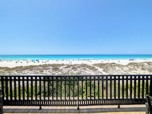 Blick auf einen Strand von einem Balkon mit Holzboden und Metallgeländer, strahlend blauer Himmel.