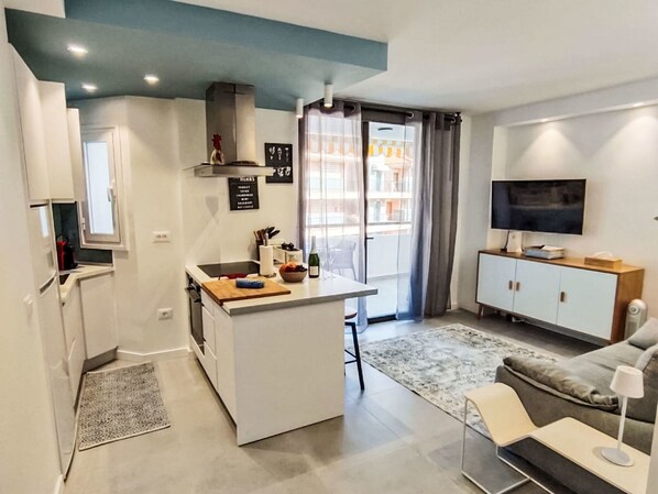 Ein kompakter Wohnbereich mit Küchenzeile, Essbereich, Sofa, Fernseher und einem Balkon mit Vorhängen.