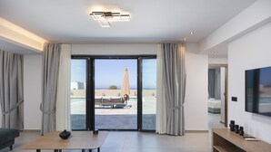 A modern hotel room with a sliding glass door to an outdoor area, a flat-screen TV, a wooden table, and a sofa. - Villa Selene – Luxury Sea View Villa (Panormo)