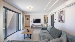 A modern living room with a sofa, a coffee table, a flat-screen TV, and a staircase leading to another level. - Villa Selene – Luxury Sea View Villa (Panormo)
