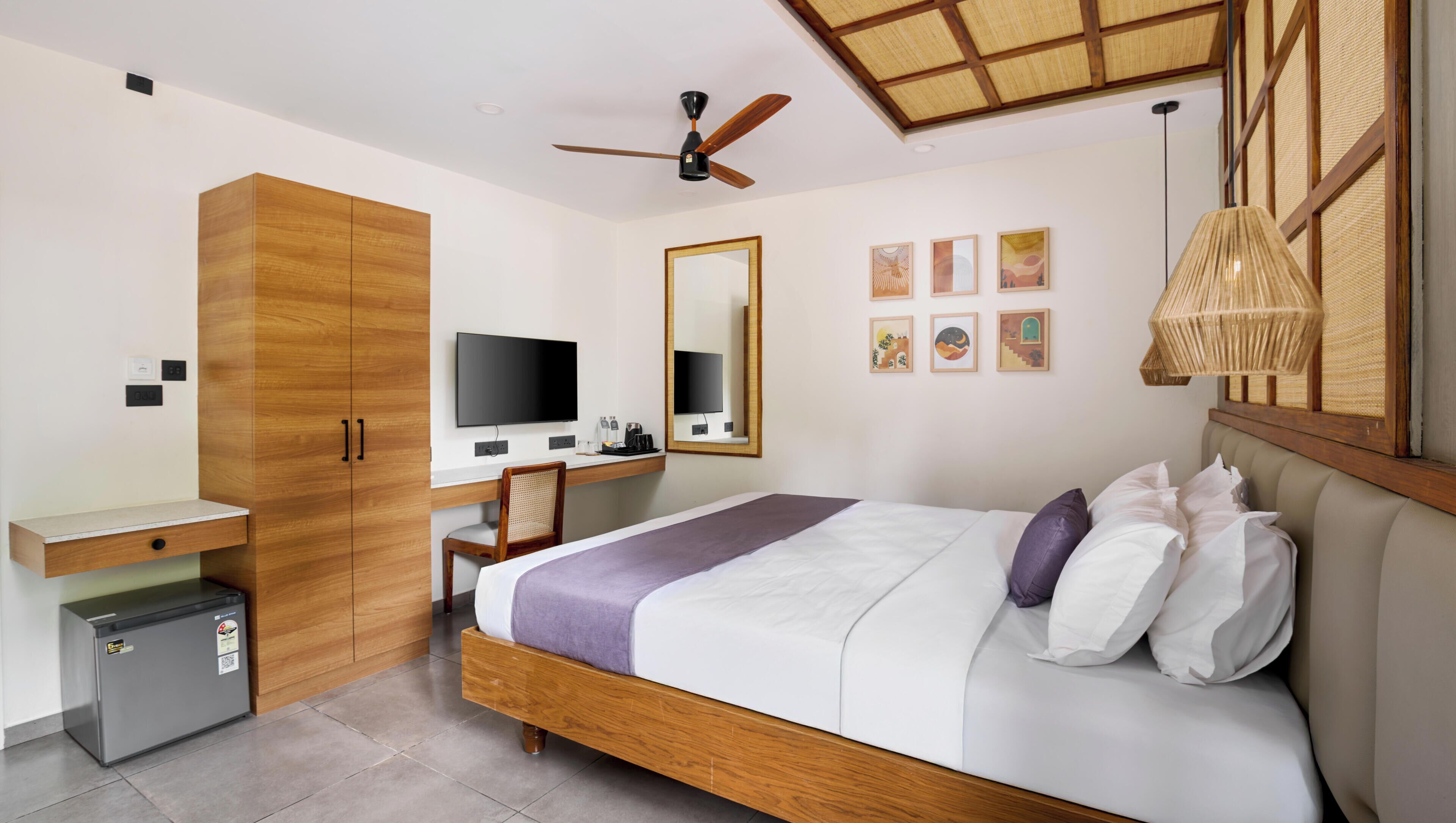 Una habitación de hotel moderna con una cama de madera, un escritorio con computadora, un ventilador de techo y un refrigerador pequeño.