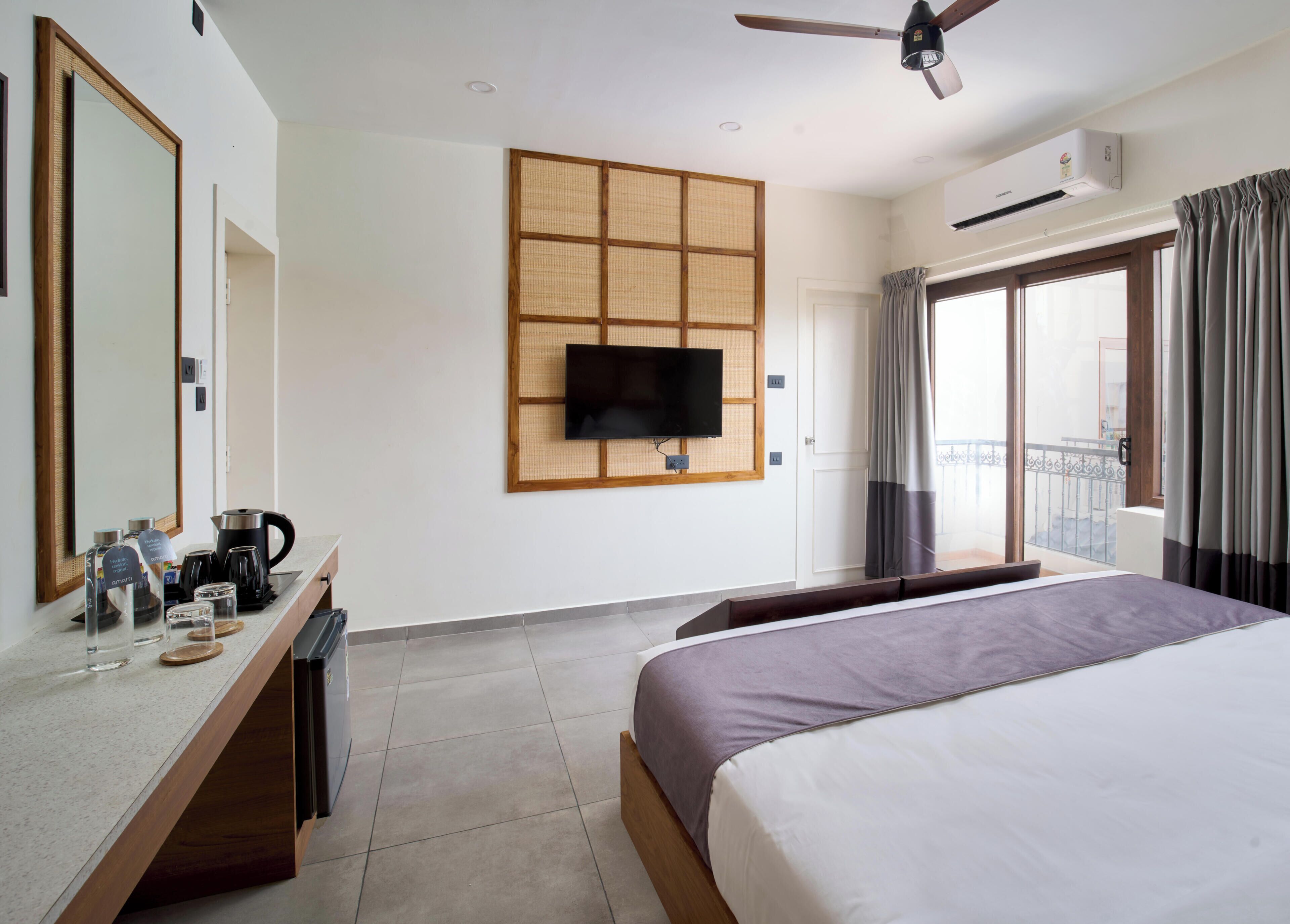 Habitación de hotel con cama, televisión montada en la pared, cafetera, hervidor de agua, un mueble pequeño y balcón con cortinas.