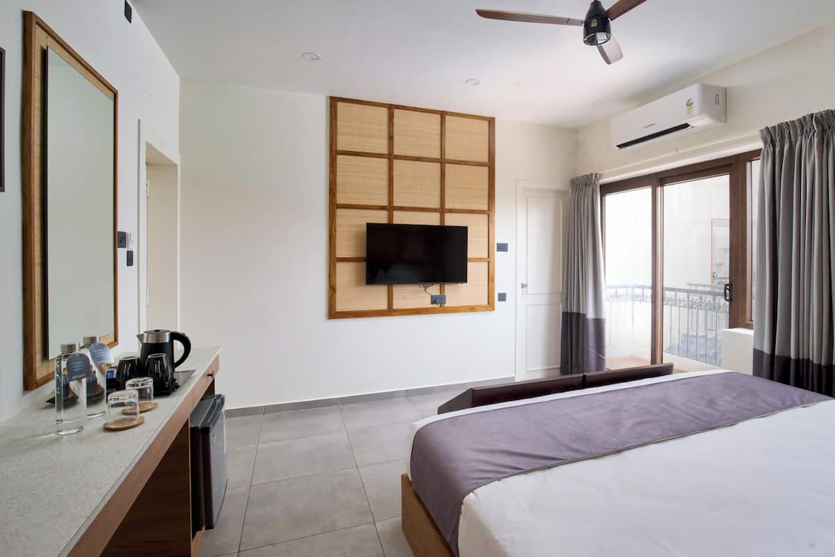 Habitación de hotel con cama, televisión montada en la pared, cafetera, hervidor de agua, un mueble pequeño y balcón con cortinas.