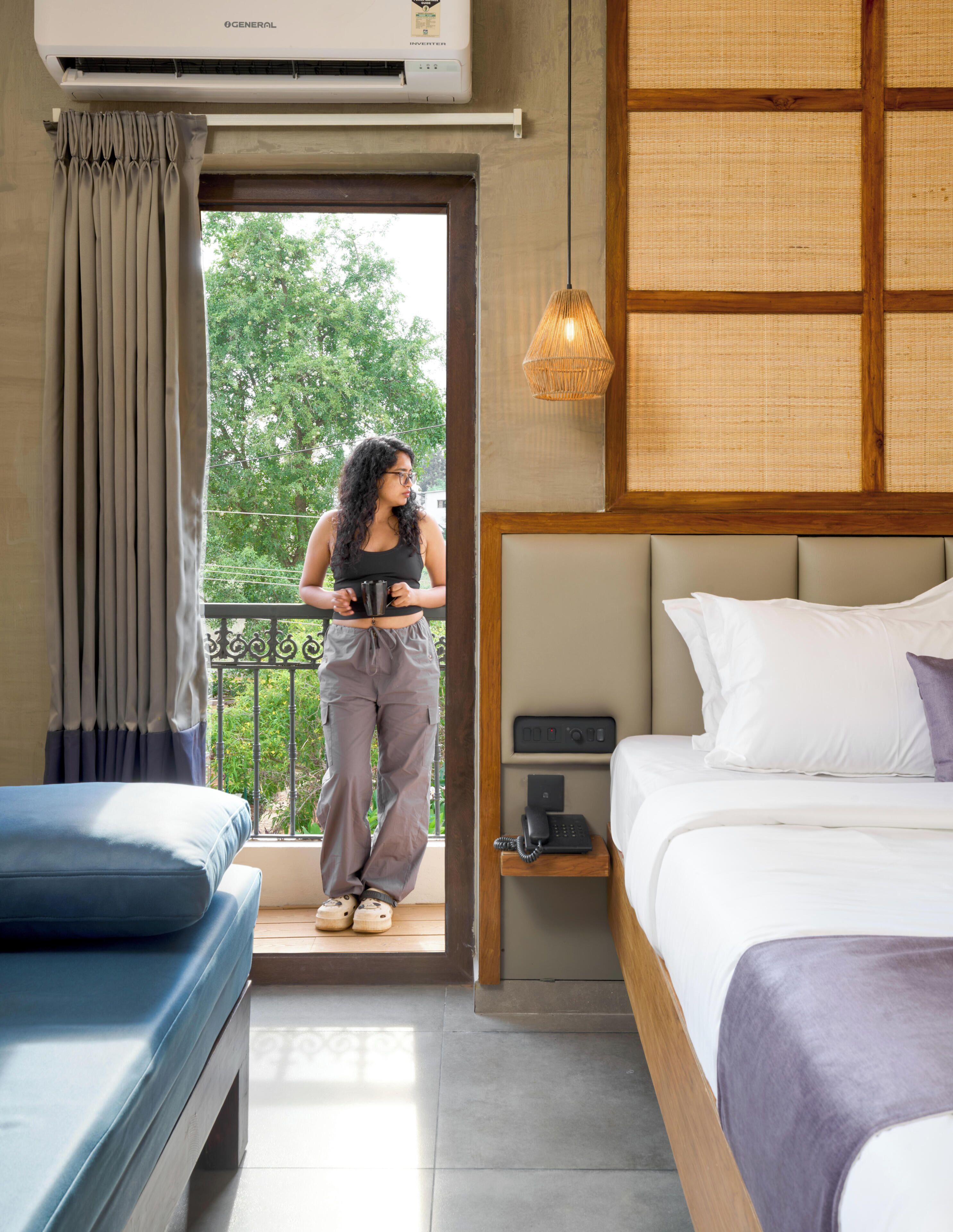 Habitación de hotel con cama, mesita de noche con teléfono, ventana con cortinas, balcón con una persona sosteniendo una taza, y un equipo de aire acondicionado.