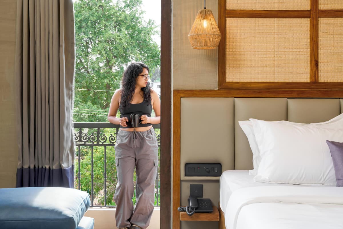 Habitación de hotel con cama, mesita de noche con teléfono, ventana con cortinas, balcón con una persona sosteniendo una taza, y un equipo de aire acondicionado.