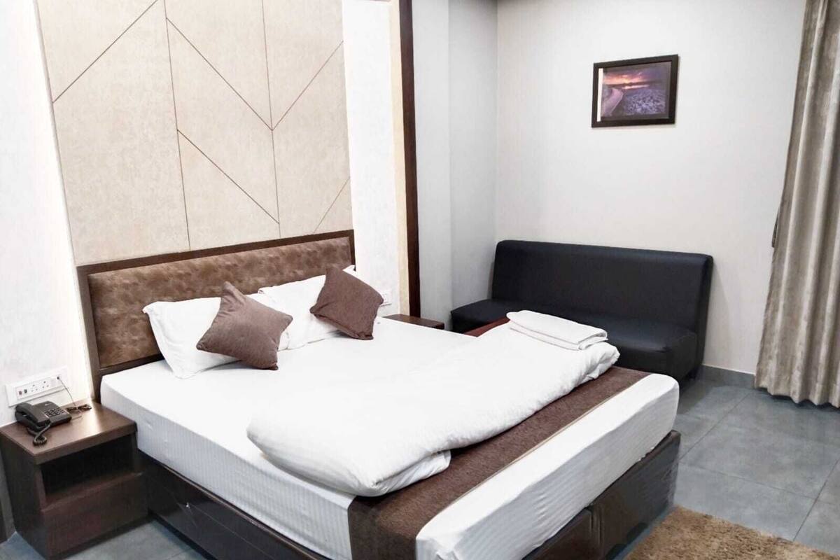 A modern hotel room with a large bed, a bedside table with a phone, a black sofa, and a framed picture on the wall.