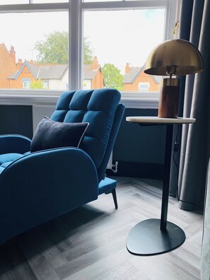Ein modernes Zimmer mit einem blauen Sofa, einem runden Tisch mit einer Lampe und Blick auf Häuser durch ein Fenster.