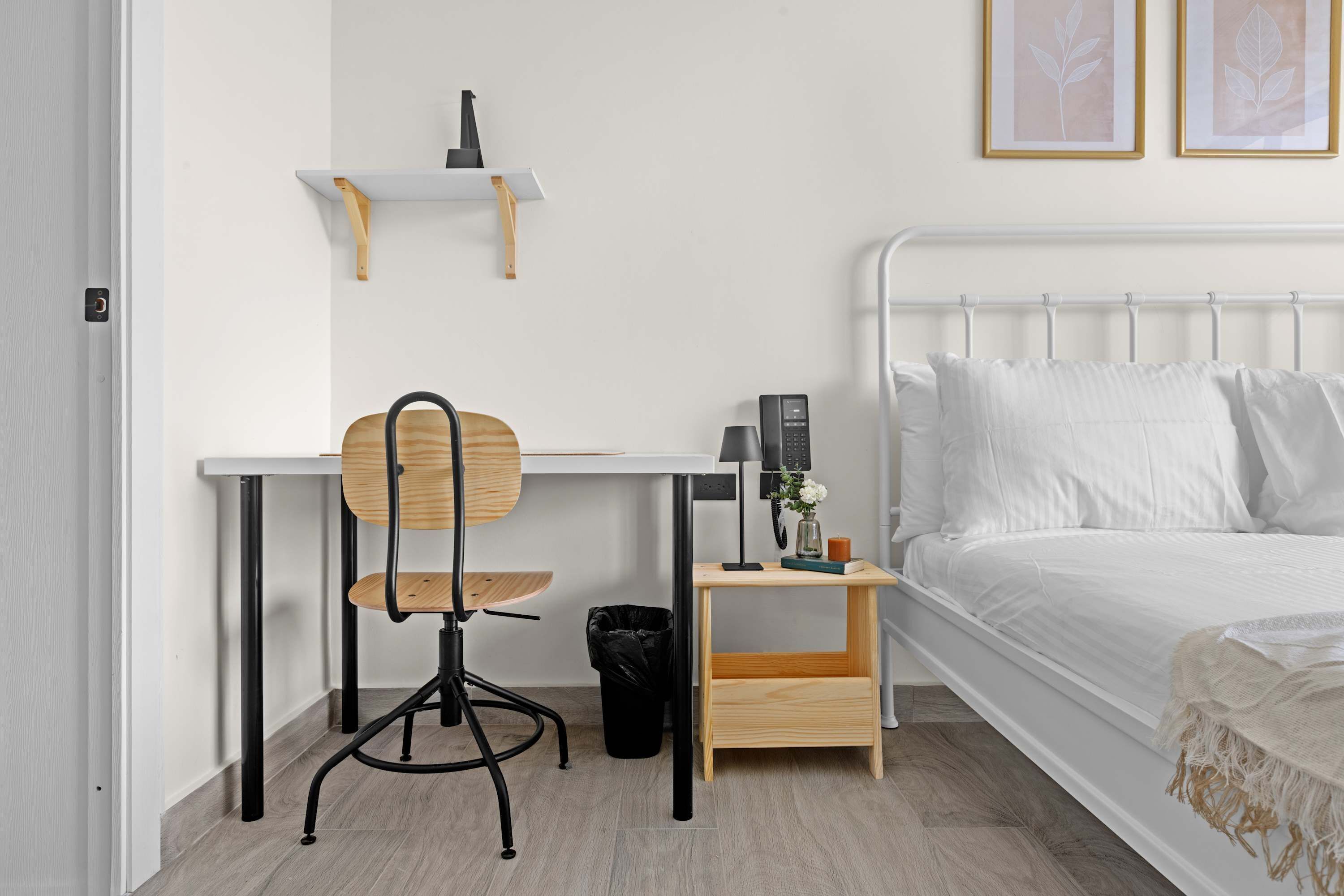 A bedroom with a bed, a desk with a chair, a small table with a lamp, and a wall-mounted shelf.