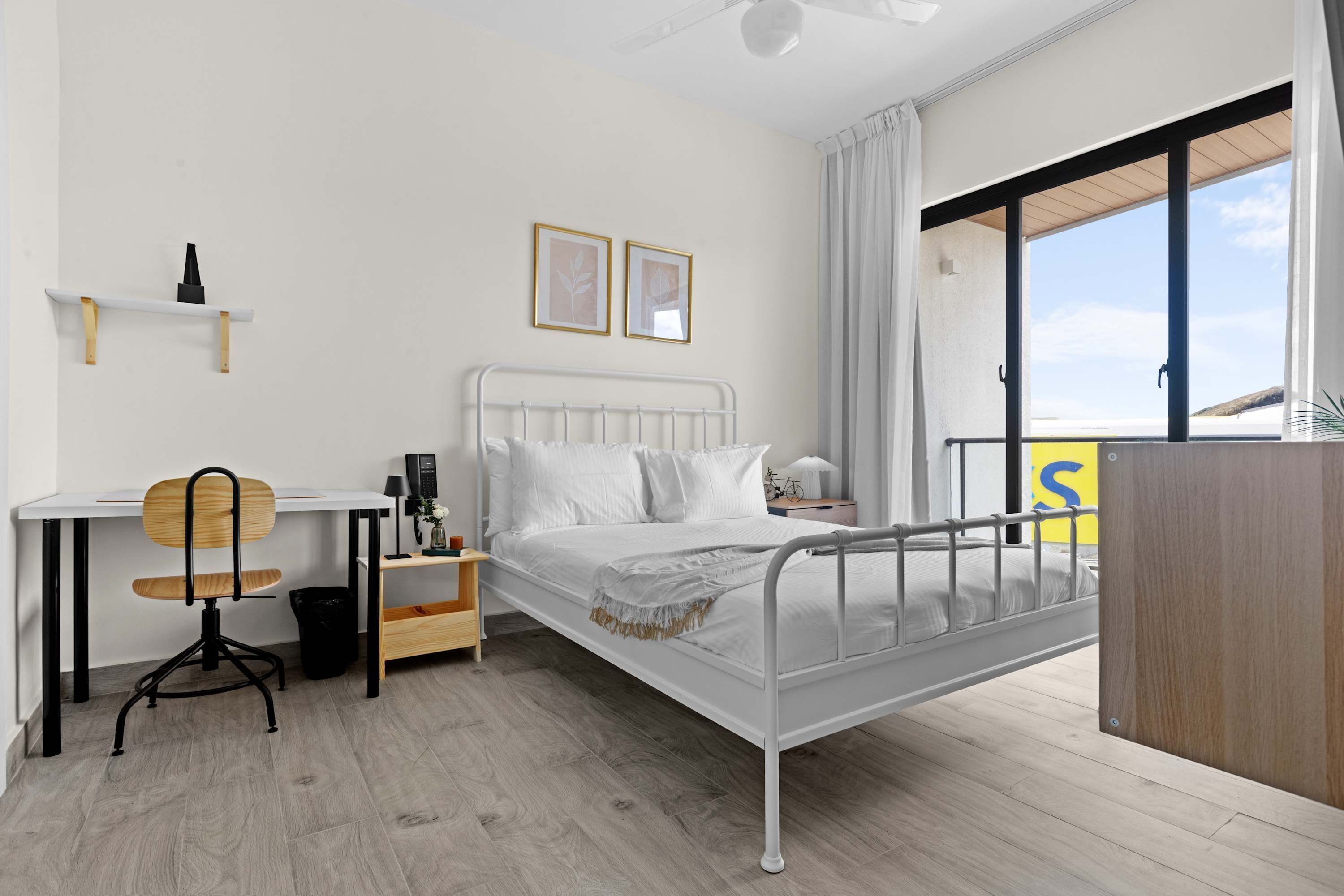 A modern bedroom with a metal bed frame, a wooden desk, a chair, a bedside table, and a view of the outdoors.