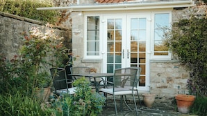 A stone building with a white door and windows, a small patio with a table and chairs, and potted plants. - Traditional Cottage Near Richmond Yorkshire Dales (Richmond)