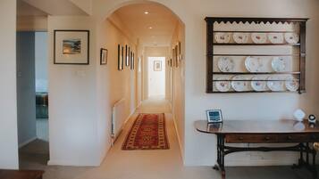 A hallway with a carpet, framed pictures on the wall, a shelf with plates, and a desk with a laptop.