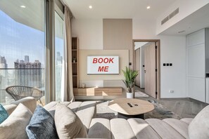 A modern living room with a large window, a sofa set, a coffee table, and a 'BOOK ME' sign on the wall. - Free Housekeeping Visits During Your Stay - Stayshort - 1 BR in Business Bay Sleeps 4 With Burj Views (Dubai)