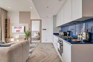 A modern kitchen with a blue countertop, white cabinets, and a built-in stove. There is a dining area with a sofa and a TV mounted on the wall. - Free Housekeeping Visits During Your Stay - Stayshort - 1 BR in Business Bay Sleeps 4 With Burj Views (Dubai)