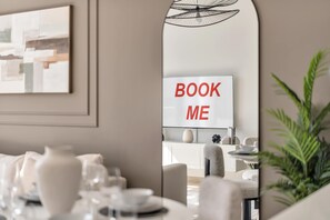 Ein modernes Interieur mit Essbereich, einem Spiegel, der das Schild 'BOOK ME' reflektiert, und einer Pflanze im Vordergrund.