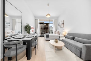 A modern living room with a dining area, grey sofa, and a large window with a city view. - Free Housekeeping Visits During Your Stay - Stayshort - Beauriful Modern Studio That Sleeps 4 at Riviera Beachfront (Dubai)