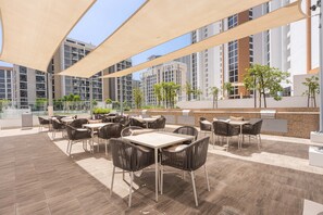 Ein Außenbereich mit Terrasse, Tischen, Stühlen, einem Grill und Blick auf hohe Gebäude.