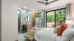 Camera da letto con un letto, una ventola a soffitto, una finestra vista verde, un comodino e un tavolino.