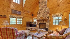 A log cabin interior with a stone fireplace, plaid furniture, a television, and a fireplace mantel.