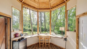 A corner room with a wooden ceiling, large windows, a small desk, two chairs, and a refrigerator. - Whidbey Island Tiny Home Near Trails & Beaches! (Oak Harbor)