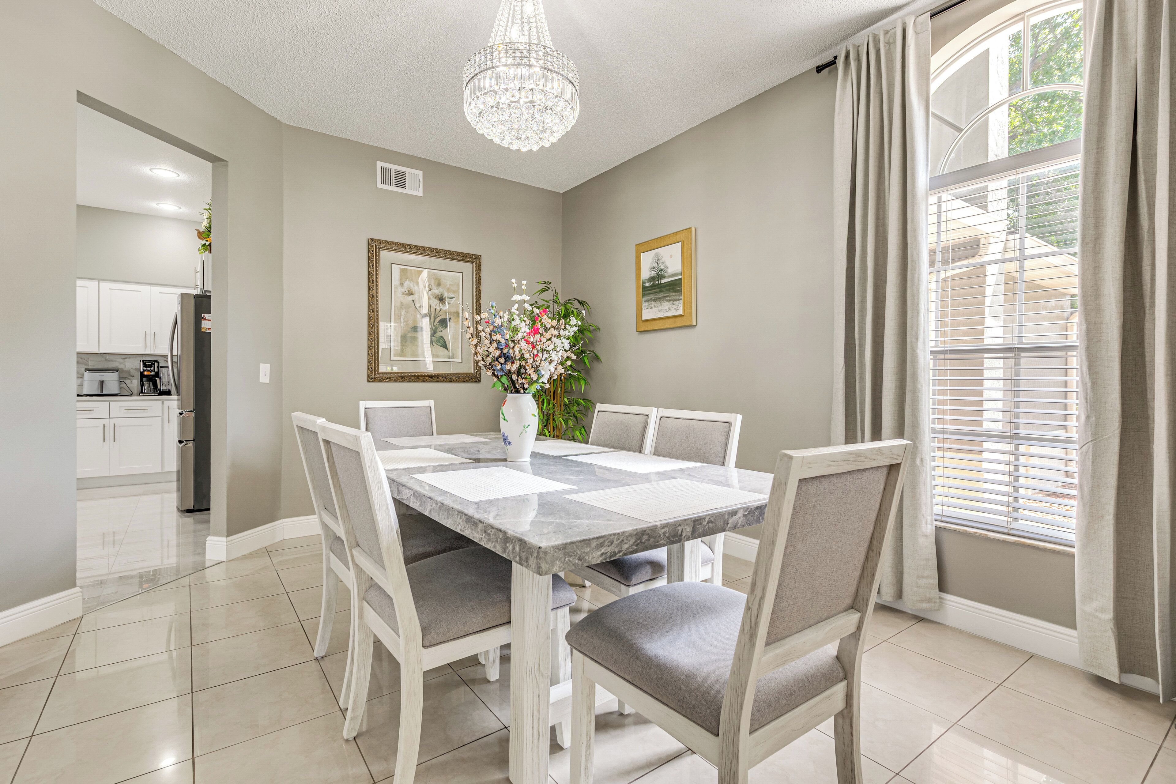 Un espace repas comprenant une table en marbre, des chaises à revêtement gris, un lustre, des œuvres d’art encadrées et une fenêtre avec des stores.
