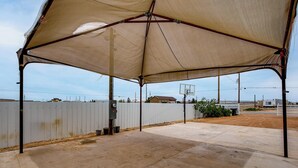 An outdoor covered area with a basketball hoop, concrete flooring, and a white fence in the background. - Family Friendly! Rural El Paso Home: Patio & Grill (El Paso)