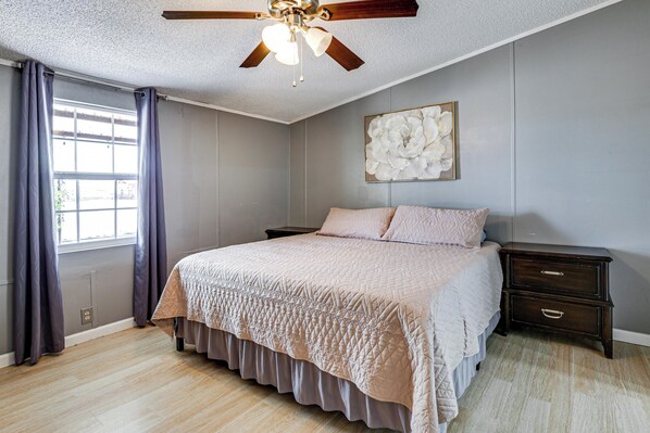 Une chambre à coucher comprenant un lit, une table de chevet, un ventilateur de plafond, une fenêtre avec des rideaux et une œuvre d’art accrochée au mur.
