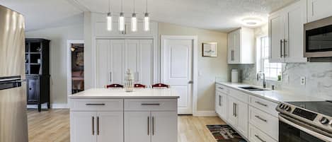 Una cocina moderna con armarios blancos, una isla central, electrodomésticos de acero inoxidable e iluminación colgante.