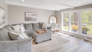 A modern living room with a sectional sofa, ottoman, and a large window with a view of greenery. - The Cottage on Lake Bernice - Campbellsport (Campbellsport)