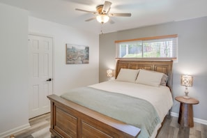 A bedroom with a bed, a ceiling fan, a window with blinds, a bedside table with a lamp, and a door.