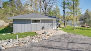 A small, single-story building with a gray exterior, a concrete foundation, and a gravel driveway leading to a wooden dock by a lake. - The Cottage on Lake Bernice - Campbellsport (Campbellsport)