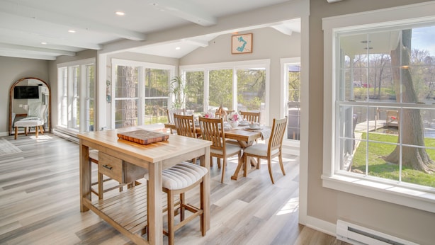 A spacious dining area with a wooden table and chairs, large windows, and a view of the outdoors. - The Cottage on Lake Bernice - Campbellsport (Campbellsport)