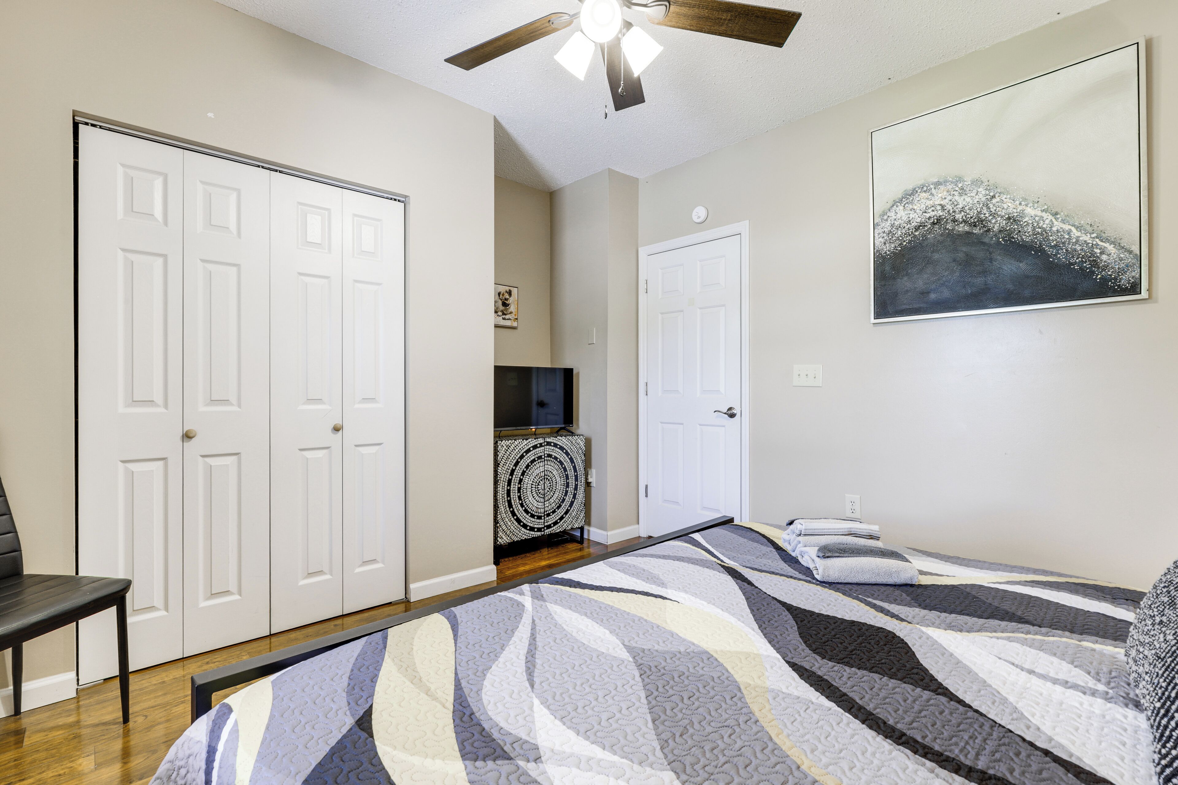 A bedroom with a bed, a ceiling fan, a door, and a wall-mounted artwork.