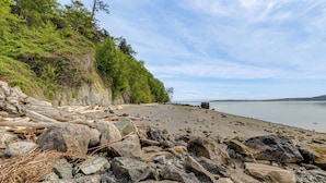 A rocky shoreline with large boulders and driftwood, a calm body of water, and a forested cliff. - Walk to Private Beach: Bay-view Whidbey Island Gem (Oak Harbor)