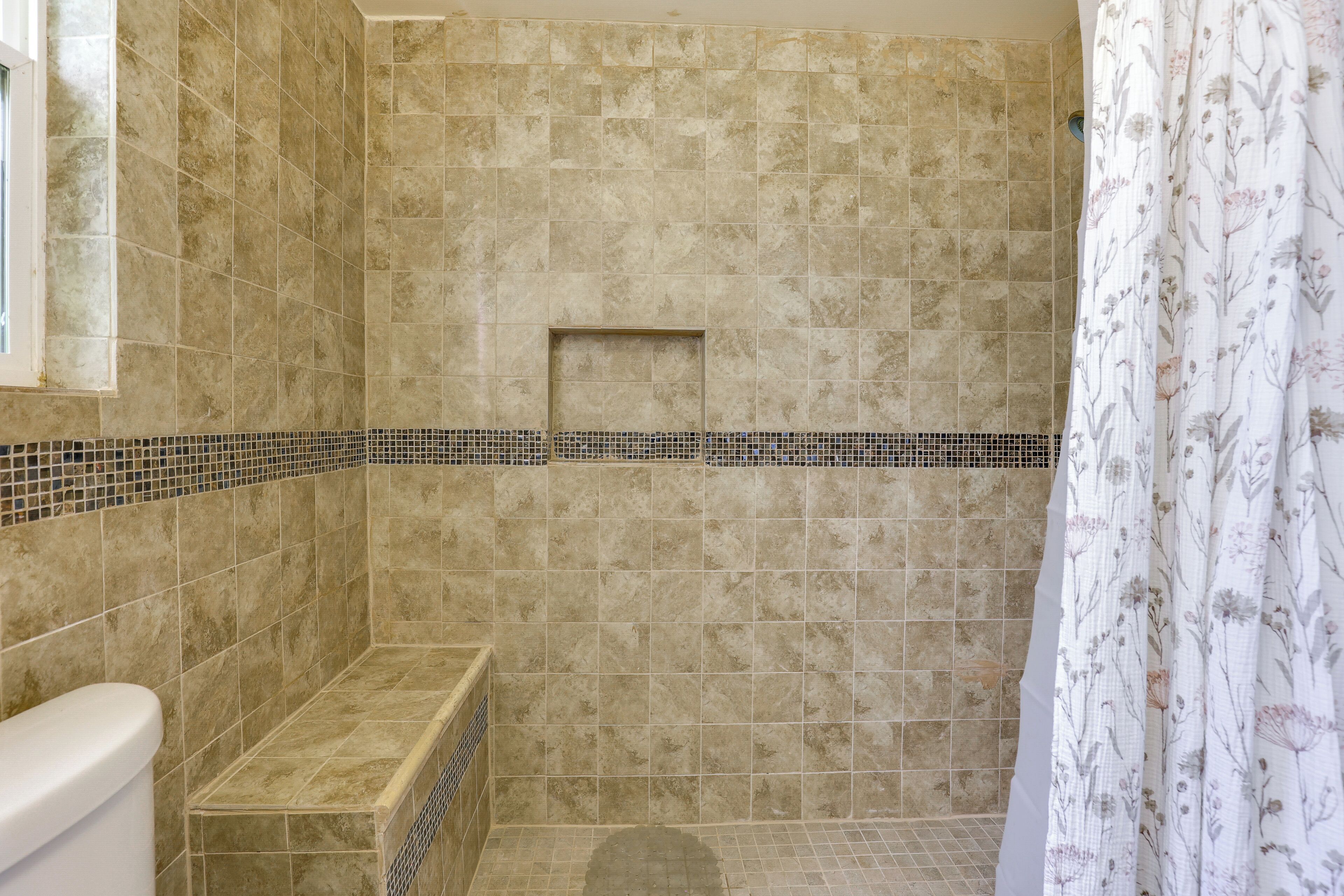 A bathroom with beige tiles, a shower area, a bench, and a window.
