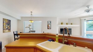 Une cuisine dotée d’un grand îlot central, d’un évier à deux bacs et d’un ventilateur de plafond. Des objets décoratifs sont disposés sur le rebord de la fenêtre, et des œuvres d’art ornent les murs.
