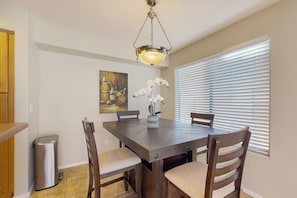 A dining area with a wooden table, four chairs, a trash can, a painting, and a window with blinds. - 2BR Condo in Fountain Hills w/ Mountain Views (Fountain Hills)