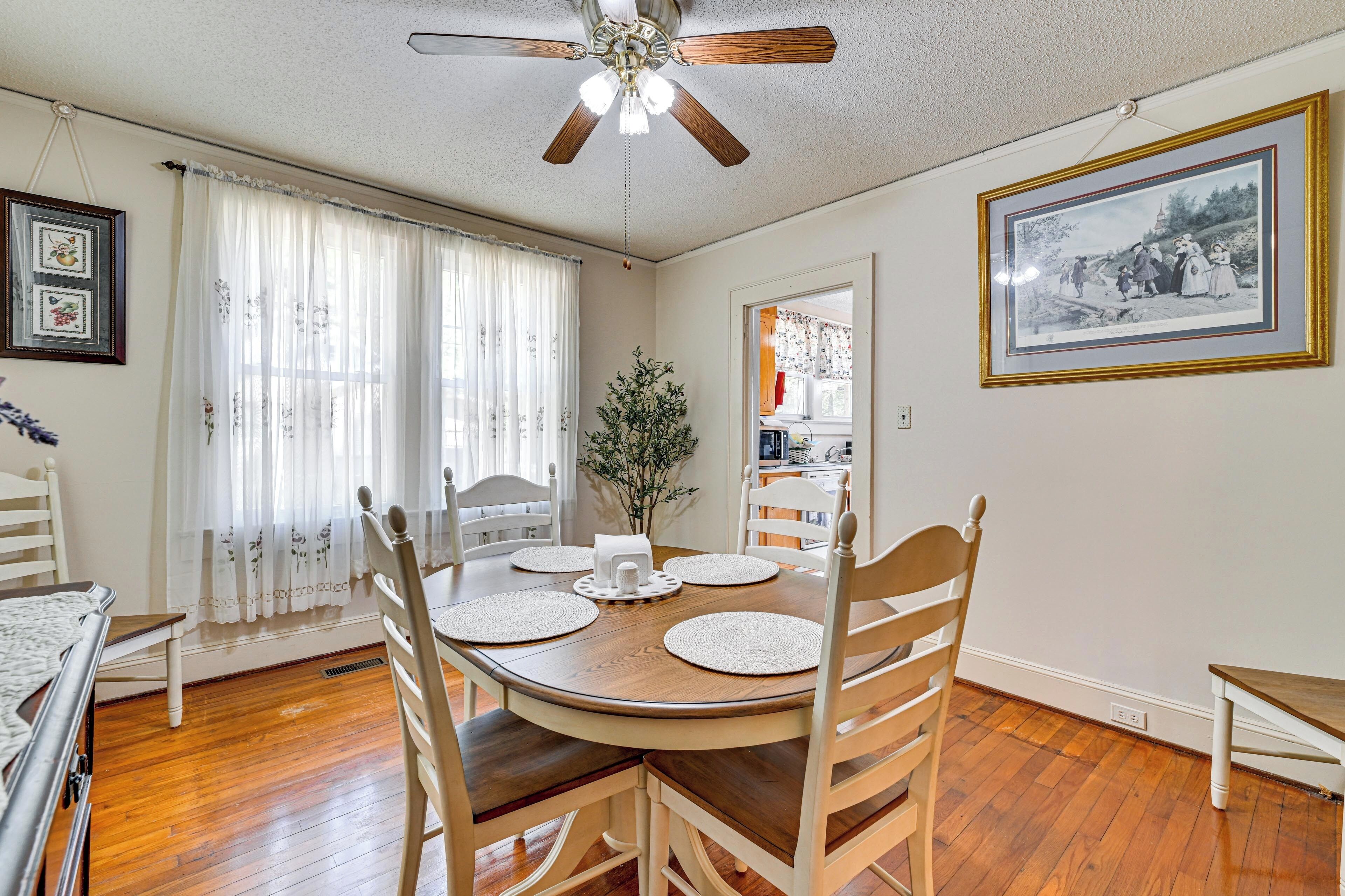 A dining room with a wooden table, white chairs, a ceiling fan, and a framed picture on the wall.