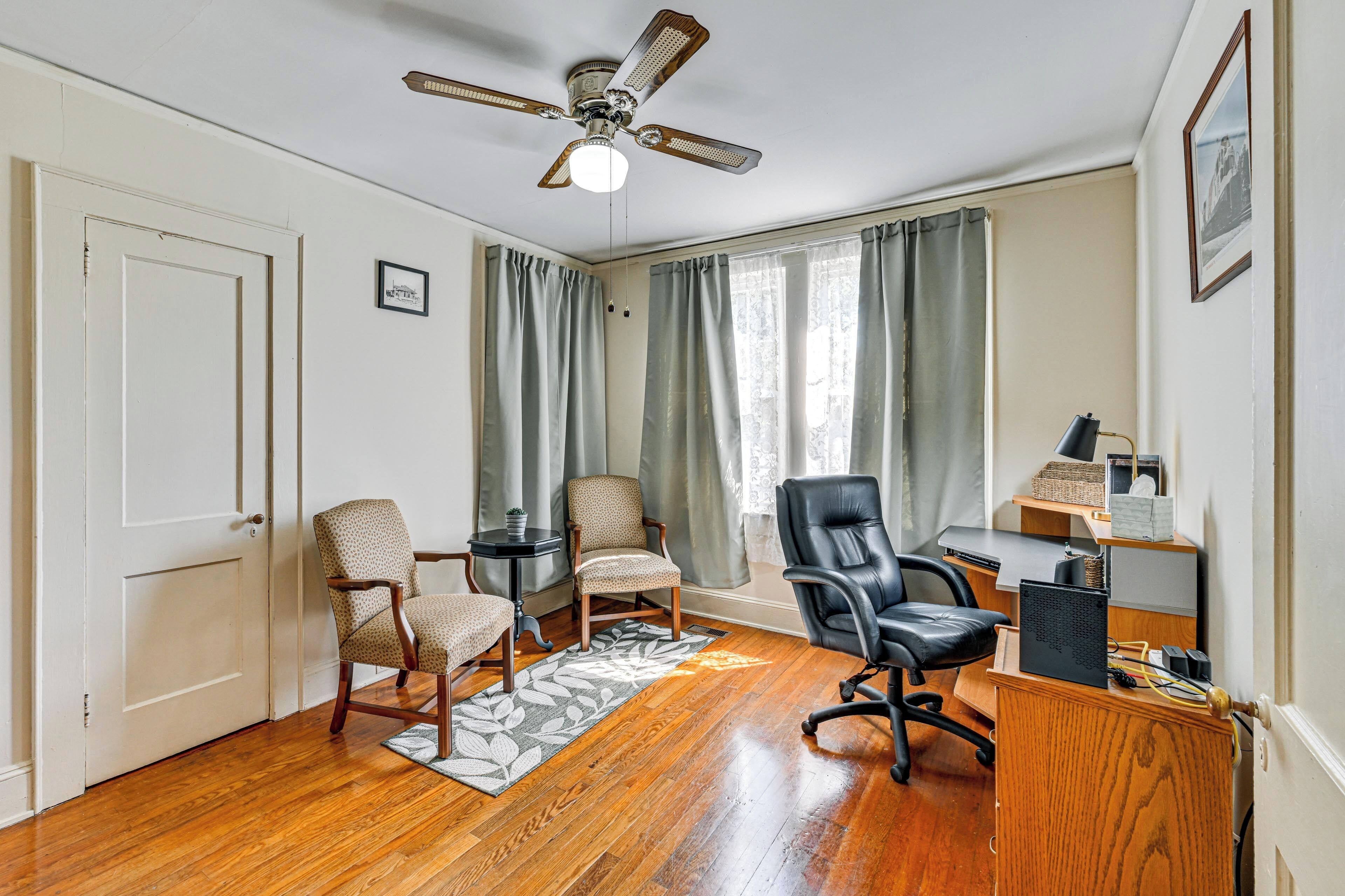 A room with hardwood flooring, a ceiling fan, two chairs, a desk with a computer, and a window with curtains.