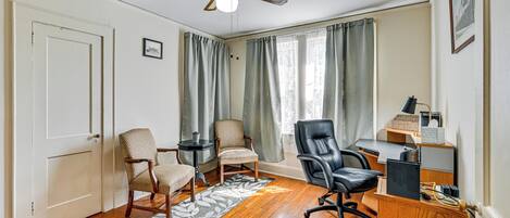 A room with hardwood flooring, a ceiling fan, two chairs, a desk with a computer, and a window with curtains.