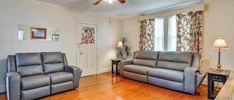 A living room with two grey sofas, a wooden floor, a ceiling fan, a door, and a window with floral curtains.
