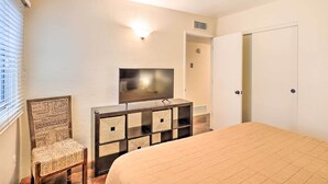 Quarto com cama, televisão sobre suporte de madeira, uma cadeira e uma porta.